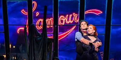 A man embraces a woman on stage in front of a large neon “L’amour” sign, with a blue-lit window and dramatic sky in the background.