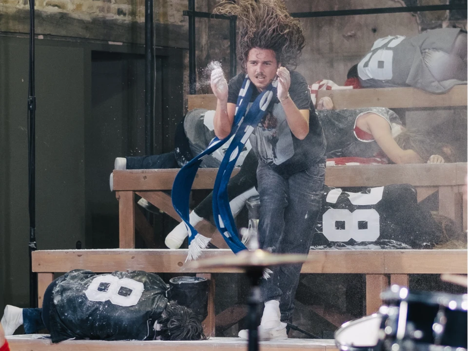 A person with long hair jumps in mid-air surrounded by others wearing numbered jerseys, some lying on wooden bleachers. White powder is visible in the air.