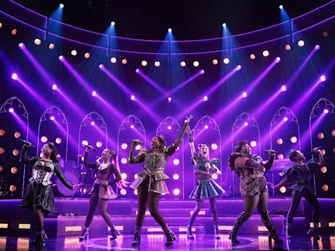 Six performers in colorful, illuminated costumes sing and pose on stage under bright purple and gold lights with an arched backdrop.