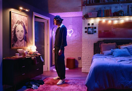 A person stands in a dimly lit bedroom with purple lighting, facing a dresser with various items and a large portrait. The bed is unmade and the walls have string lights and decorations.