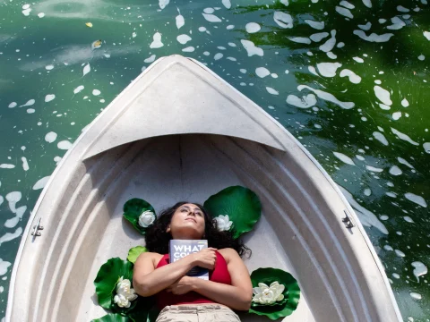 Person lying in a boat on green water, surrounded by large leaves and white flowers, holding a book titled "WHAT COULD.