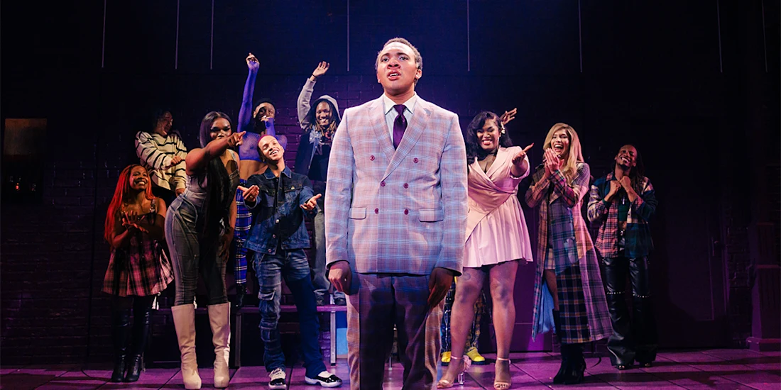 A person in a plaid suit stands center stage under a spotlight, while a group of people behind them cheer and clap with colorful outfits and expressions of excitement.