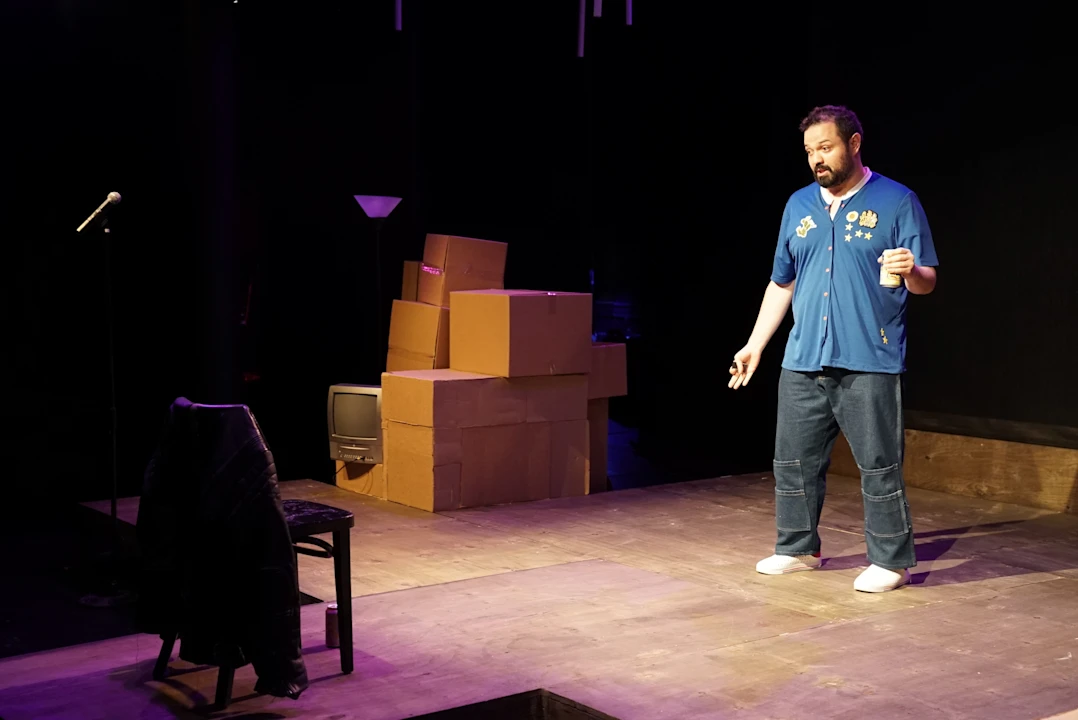 A man stands on a stage near a chair, microphone, and stacked cardboard boxes, holding an object in one hand and wearing a blue shirt and jeans.