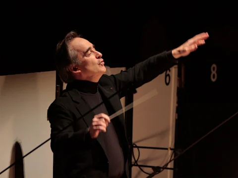 A conductor in a black suit raises one hand while holding a baton, appearing to lead an orchestra during a performance.