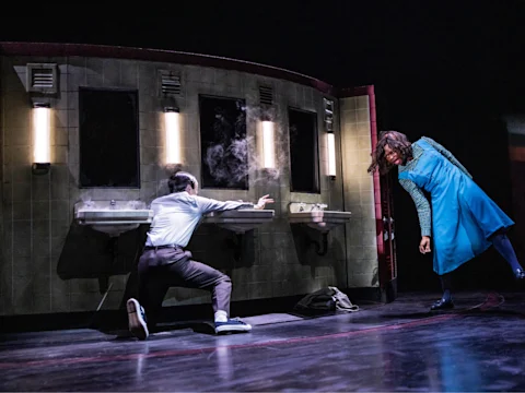 Two people perform on stage in a dramatic scene set in a bathroom with mirrors and sinks; one person crouches and reaches out as smoke rises, while the other leans back in a blue dress.