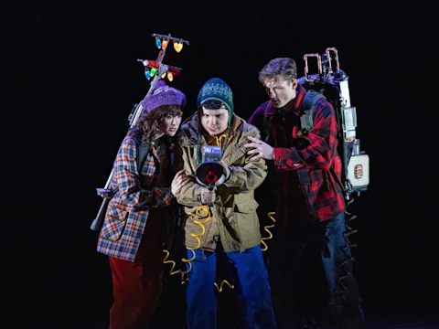 Three people in outdoor clothing and winter hats stand together, holding a large device with wires and lights, against a dark background.