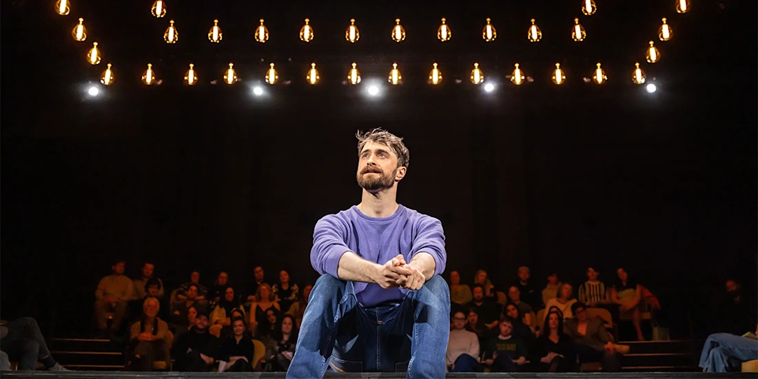 A man in a purple shirt sits cross-legged on stage under hanging lights, with an audience seated in the background.