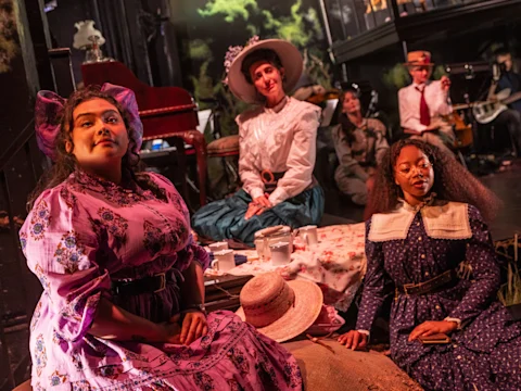 Three women in vintage dresses sit at a picnic setup on stage, while musicians and another person in period attire are visible in the background.