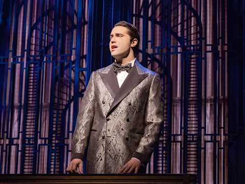 A man in a patterned suit and bow tie stands on stage in front of a decorative backdrop, appearing to sing or speak during a theater performance.