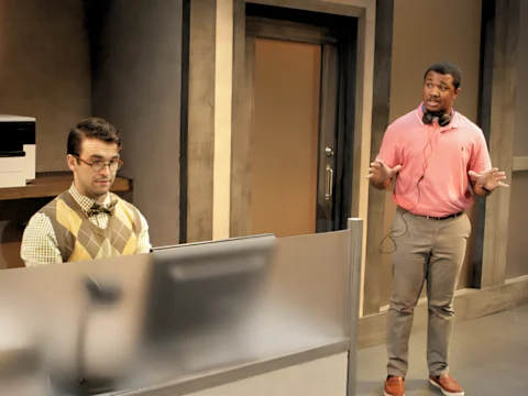 Two men in an office setting; one is seated at a computer wearing glasses and a bow tie, while the other stands nearby gesturing with hands, wearing a pink shirt and headphones around his neck.