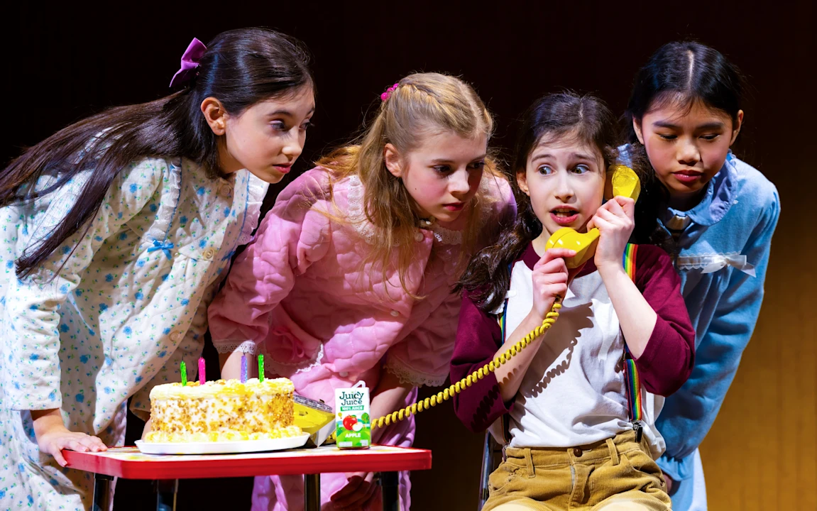 Four girls gather around a table with a cake. One girl is speaking on a yellow phone while the others listen attentively.