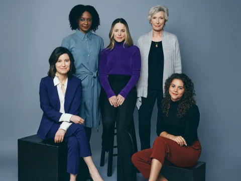 A group of five women pose against a plain background. Three are standing, and two are seated. They are dressed in business-casual attire.