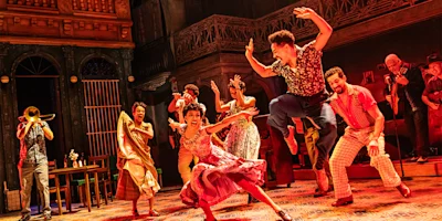 A group of performers in colorful costumes dance energetically onstage, with musicians playing instruments in the background, set against a vibrant, urban backdrop.