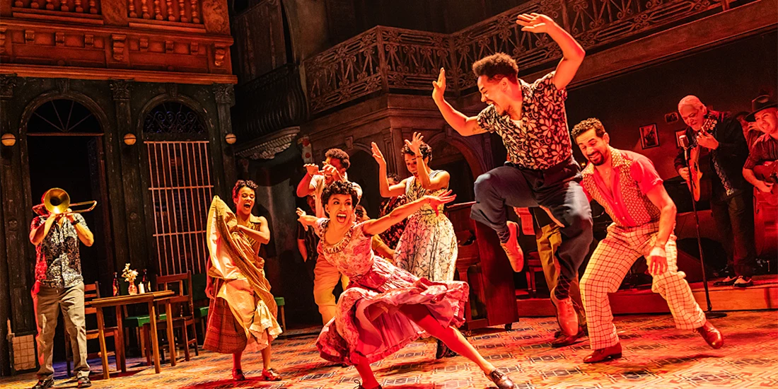 A group of performers in colorful costumes dance energetically onstage, with musicians playing instruments in the background, set against a vibrant, urban backdrop.