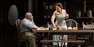Two people in vintage-style clothing sit and stand at a wooden dining table set for a meal in a dimly lit kitchen with antique furniture and dishes.