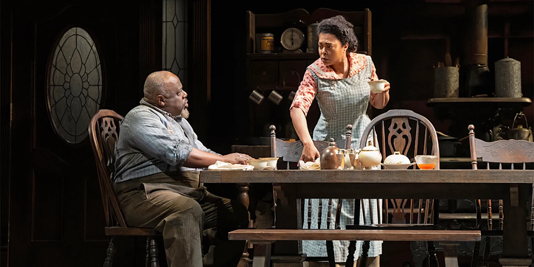 Two people in vintage-style clothing sit and stand at a wooden dining table set for a meal in a dimly lit kitchen with antique furniture and dishes.