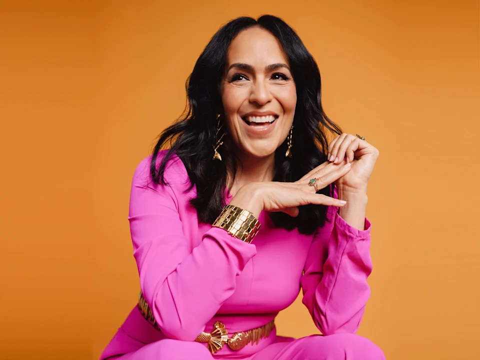 A woman with long dark hair, wearing a bright pink outfit and gold accessories, smiles against an orange background.