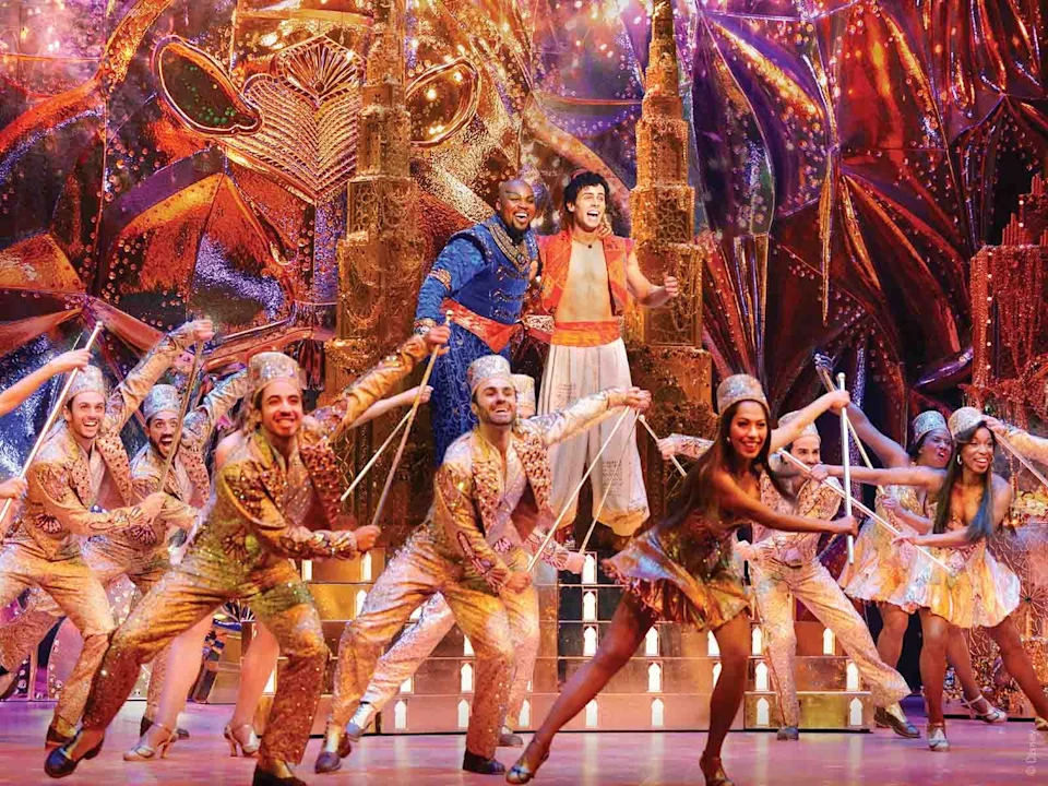 A stage performance of "Aladdin" with actors in colorful costumes dancing, featuring Aladdin and Genie in the center against a vibrant, illuminated backdrop.