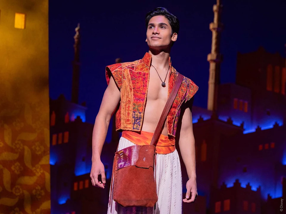A man in a colorful vest, white pants, and a brown satchel stands on stage with a backdrop of illuminated buildings and towers.