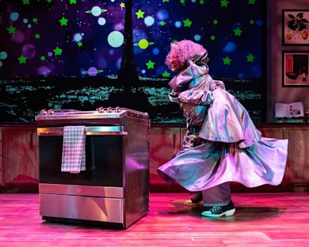 A person in a shiny silver coat and green sneakers stands by a stove on a stage with a space-themed backdrop and wooden cabinets.