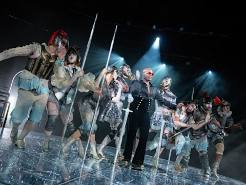 A group of performers in elaborate, futuristic costumes pose on stage under dramatic lighting, holding long white poles.