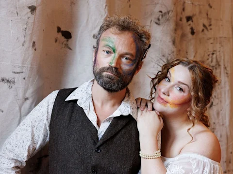 A man and woman with colorful face paint stand together in front of a textured fabric backdrop, both looking at the camera.