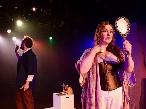 A woman in costume holds a mirror on stage, while a man in period clothing stands in the background under theatrical lighting.