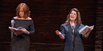 Two women stand at microphones on stage; one reads from a book while the other gestures and smiles, both in formal attire against a brick wall backdrop.