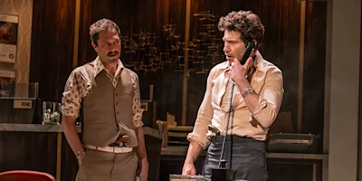 Two men in 1970s-style clothing stand in an office; one is talking on a corded phone while the other looks on.