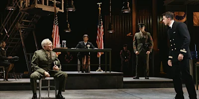 A scene from a play showing military personnel in a courtroom setting, with a seated officer speaking to a standing officer in a navy uniform while a judge presides at a desk.