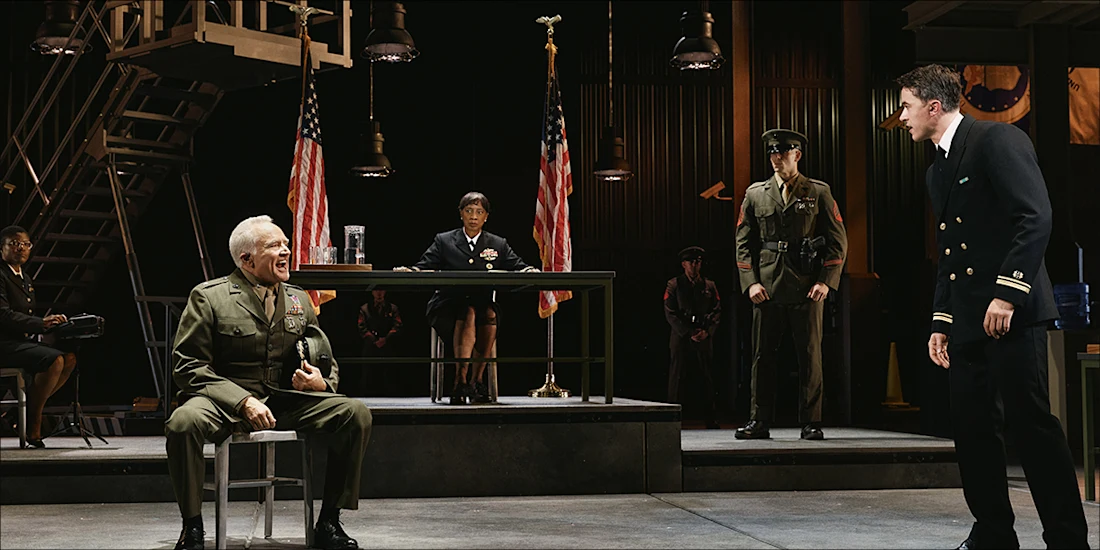 A scene from a play showing military personnel in a courtroom setting, with a seated officer speaking to a standing officer in a navy uniform while a judge presides at a desk.