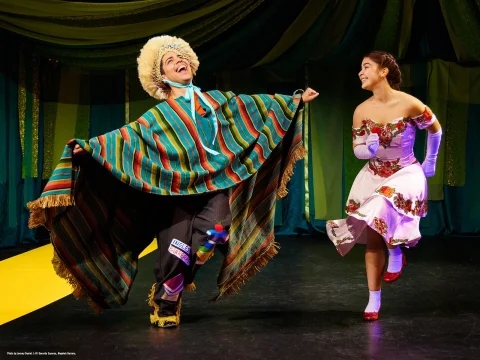 Two people perform a lively dance on stage; the person on the left wears a colorful traditional outfit and hat, while the person on the right is dressed in a floral-patterned dress and gloves.