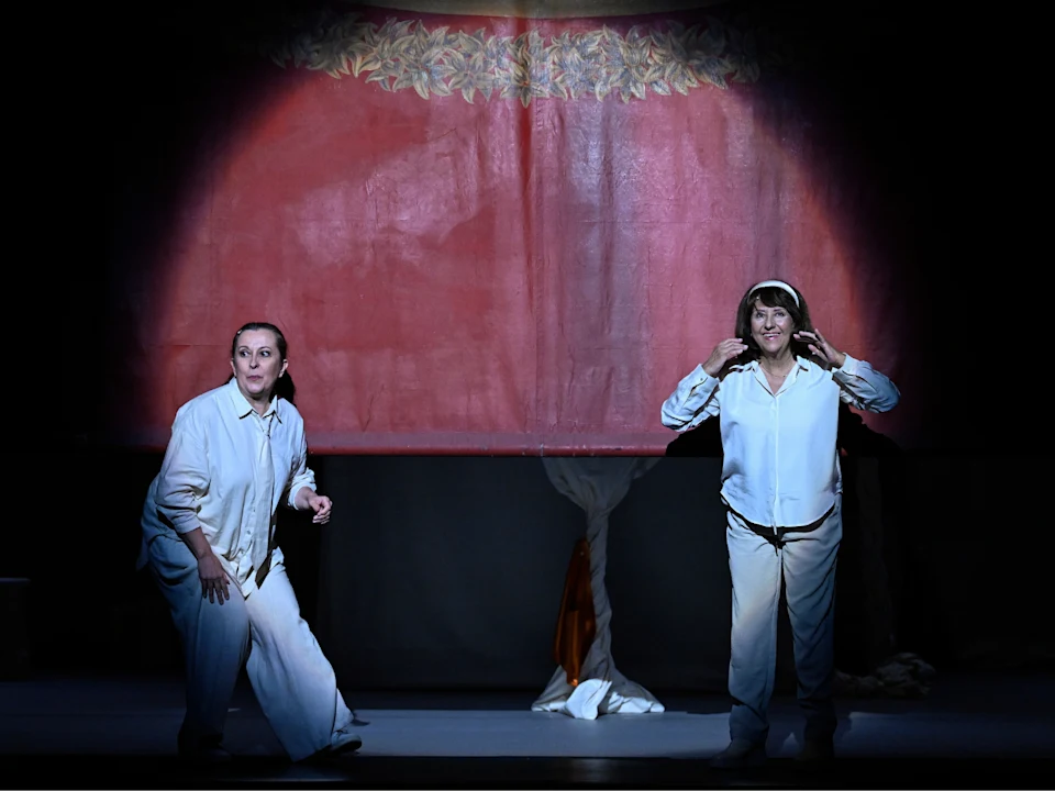 Two people in white clothing stand on stage under a spotlight, in front of a red curtain with a decorative border.