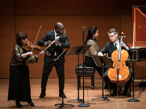 Four musicians perform on stage with a violin, flute, harpsichord, and cello. Music stands and microphones are placed in front of them against a wooden backdrop.