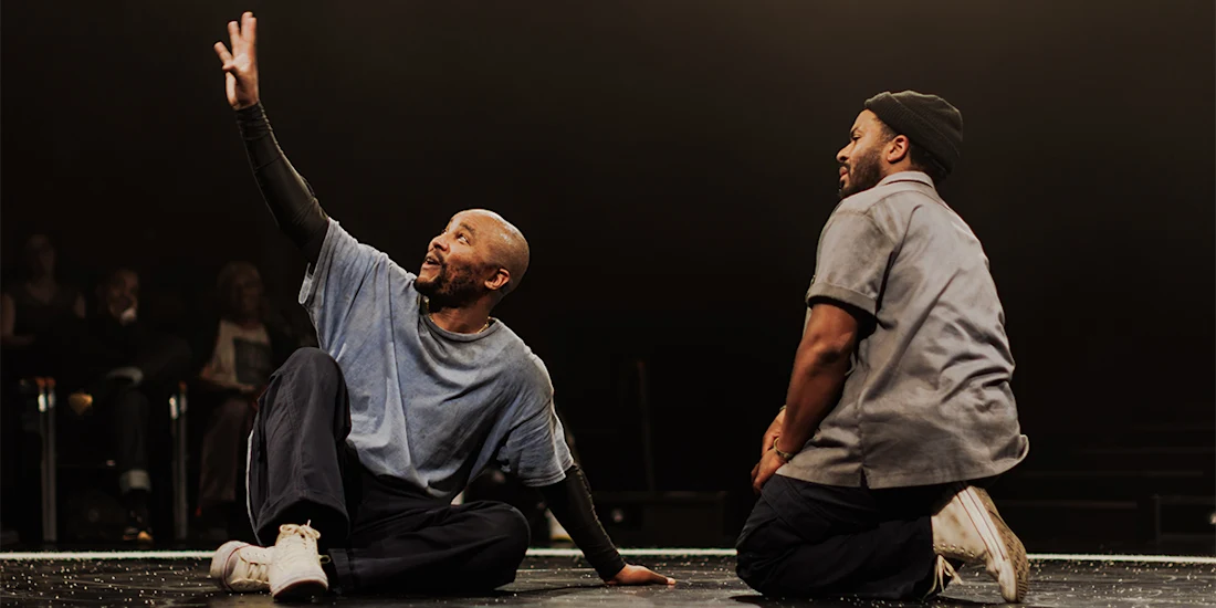 Two men kneel on stage under dramatic lighting; one gestures upward with his arm while the other looks on. Audience members are visible in the background.