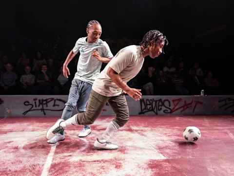 Two people in sportswear compete for a soccer ball on a graffiti-covered indoor field, with spectators watching in the background.