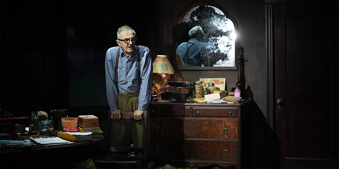 An elderly man in glasses and suspenders stands in a dimly lit, cluttered room beside a wooden dresser with a cracked mirror reflecting his image.