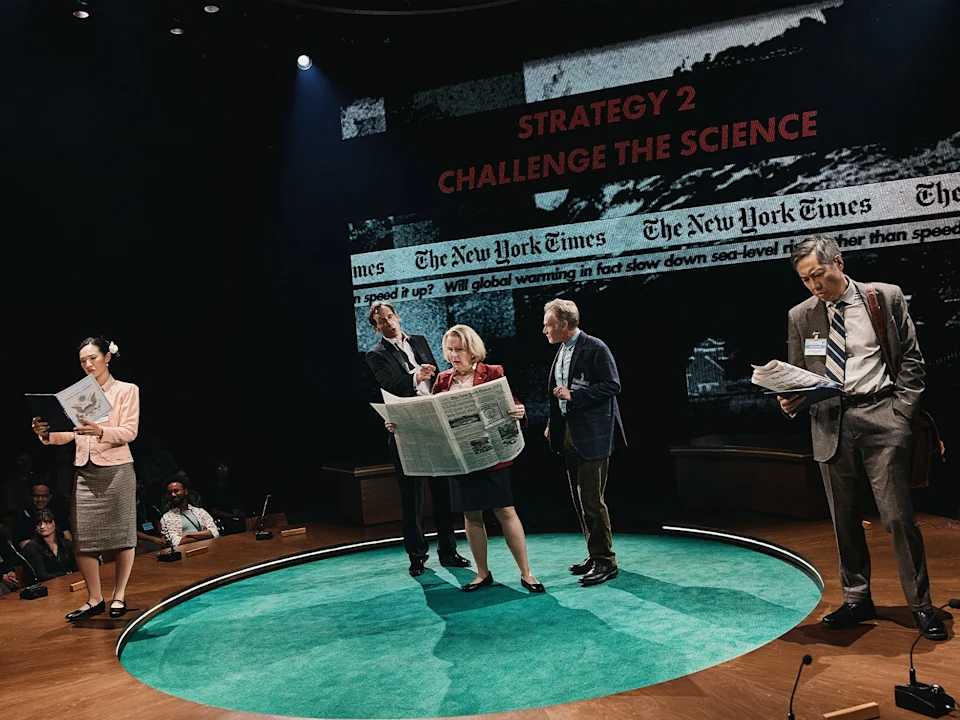 Production photo of Kyoto in New York, showing five people stand on a stage set with green carpet, holding newspapers and papers, with a projection behind them that reads "STRATEGY 2 CHALLENGE THE SCIENCE.