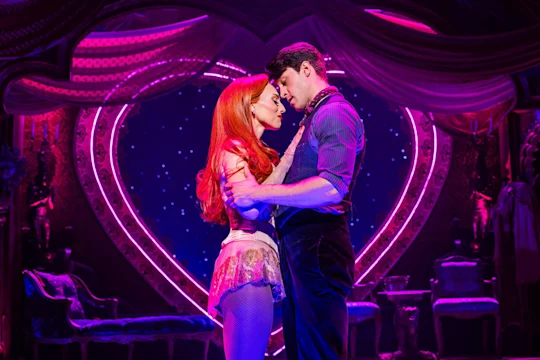 Two actors in costume embrace closely on stage, with dramatic pink lighting and a large illuminated heart shape in the background, suggesting a romantic scene in a theatrical production.