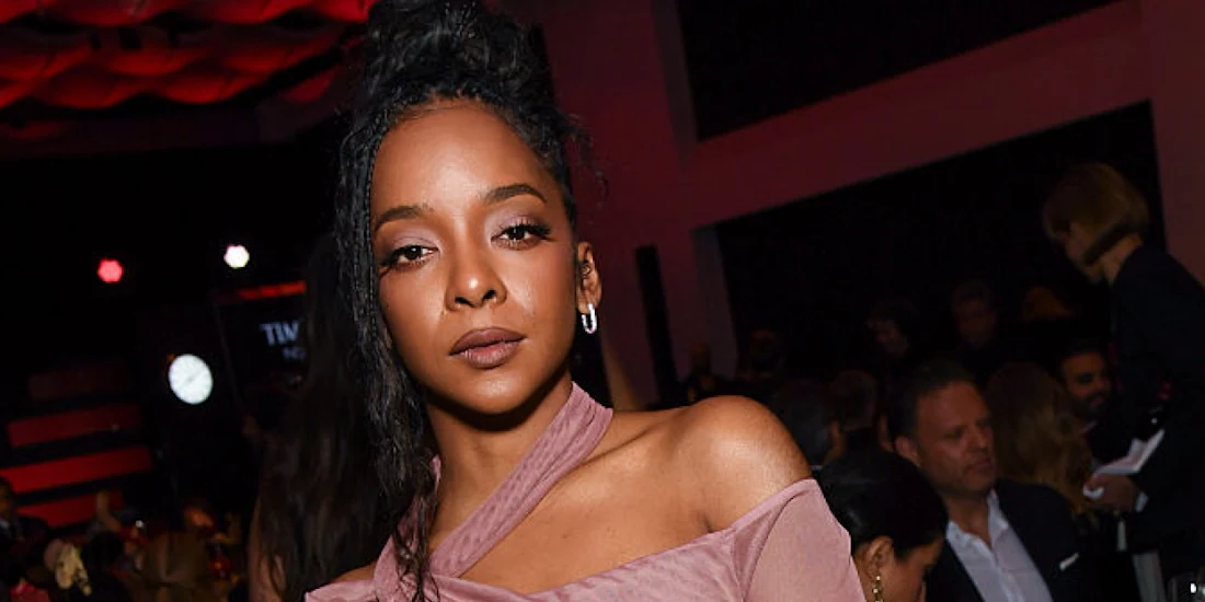 A woman in a pink off-the-shoulder top poses for a photo at an indoor event with people seated and mingling in the background.