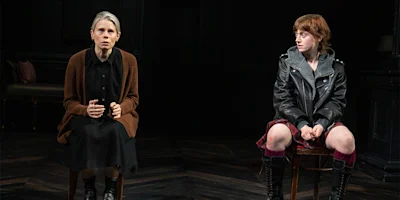 Two women sit on chairs in a dimly lit room; one wears a brown cardigan and black dress, appearing tense, while the other wears a black leather jacket and looks to the side.