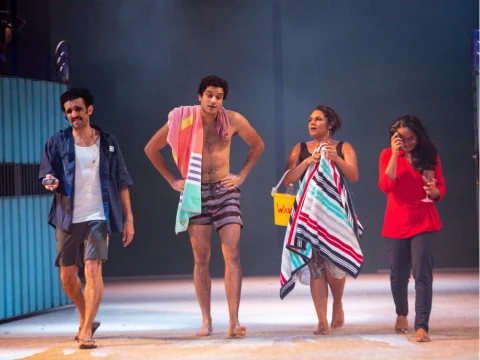 Four individuals walk barefoot on a stage with towels, beachwear, and a bucket labeled "WAX." The background suggests a beach or coastal setting with mist.