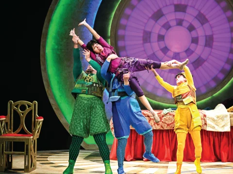 Performers in colorful costumes engage in a dramatic lift on stage, in front of a large circular backdrop, next to an empty chair and a red table.