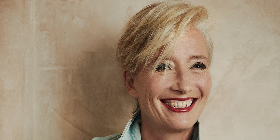 A person with short blond hair, red lipstick, and a light blue collared shirt smiles while standing against a beige textured wall.