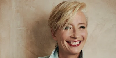 A person with short blond hair, red lipstick, and a light blue collared shirt smiles while standing against a beige textured wall.
