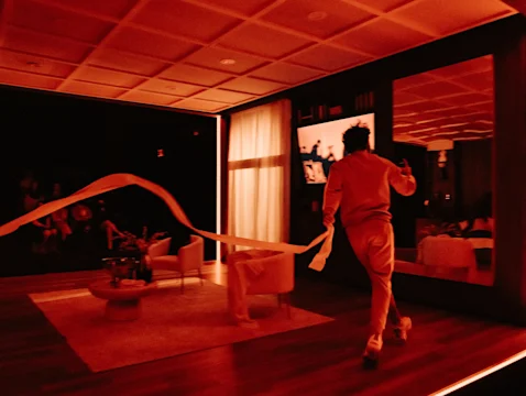 Person in dim red-lit room runs with a long roll of paper trailing behind them; modern furniture and large mirror visible.