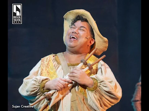 A performer in a period costume and straw hat smiles while holding a wooden prop on stage under theatrical lighting.