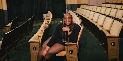 A person with long braids and glasses sits smiling in an empty theater, surrounded by rows of beige seats with green carpeting.
