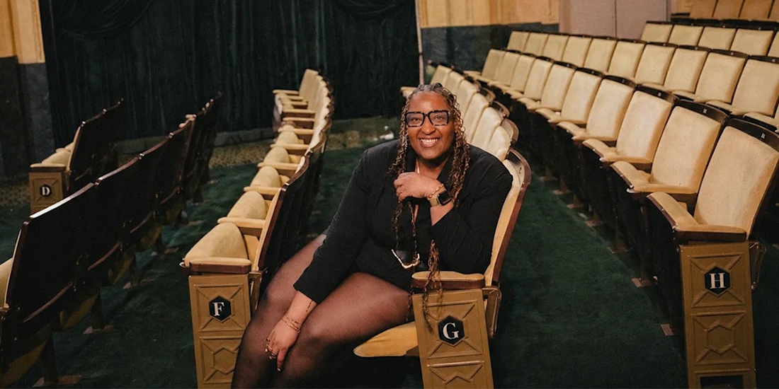 A person with long braids and glasses sits smiling in an empty theater, surrounded by rows of beige seats with green carpeting.