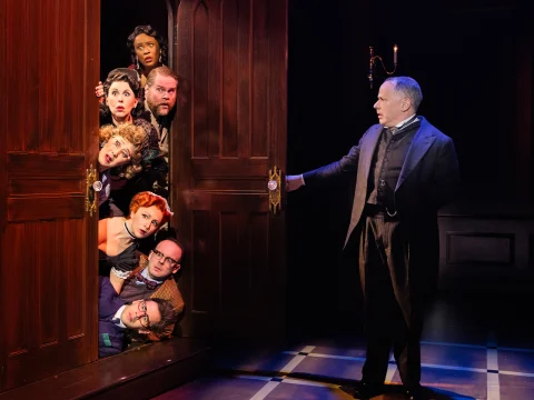 Production shot of Clue in Washington DC, showing ensemble in colorful costumes peek from behind double doors while a man in formal attire looks at them from the side. 